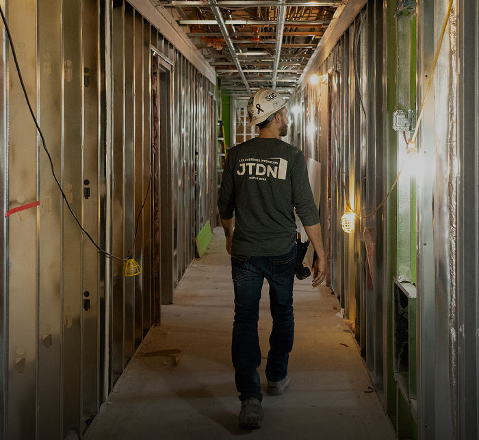 Ouvrier de JTDN marchant dans un corridor en chantier, portant un casque de sécurité et un chandail de l’entreprise, entouré de structures métalliques et de câblage en installation.